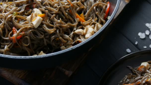 Große Nudelpfanne neben asiatischen Buchweizennudeln Soba mit Gemüse Zwiebelschoten, Karotten, Paprika und Sesam und Hühnerstücken, auf einem stilvollen schwarzen Teller und chinesischen Essstäbchen — Stockvideo