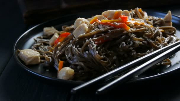 Macarrão de trigo sarraceno asiático soba com legumes vagens de cebola, cenouras, sementes de páprica e gergelim e pedaços de frango, em uma placa preta elegante e pauzinhos chineses no estilo de chave baixa — Vídeo de Stock