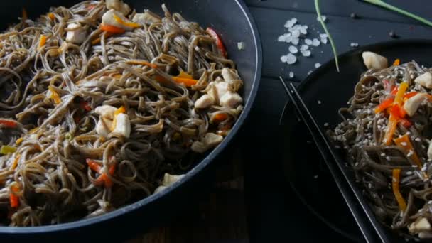 Sartén grande de fideos junto a fideos asiáticos de trigo sarraceno soba con verduras vainas de cebolla, zanahorias, pimentón y semillas de sésamo y trozos de pollo, en un elegante plato negro y palillos chinos — Vídeo de stock