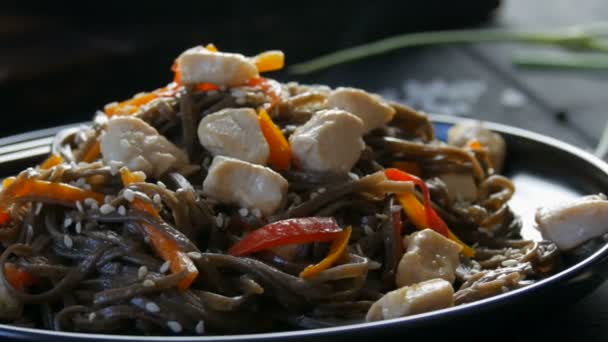 Asiatische Buchweizen-Nudeln Soba mit Gemüse Zwiebelschoten, Karotten, Paprika und Sesam und Hühnerstücken, auf einem stilvollen schwarzen Teller und chinesischen Essstäbchen im Stil der zurückhaltenden — Stockvideo