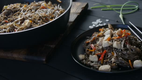 Große Nudelpfanne neben asiatischen Buchweizennudeln Soba mit Gemüse Zwiebelschoten, Karotten, Paprika und Sesam und Hühnerstücken, auf einem stilvollen schwarzen Teller und chinesischen Essstäbchen — Stockvideo