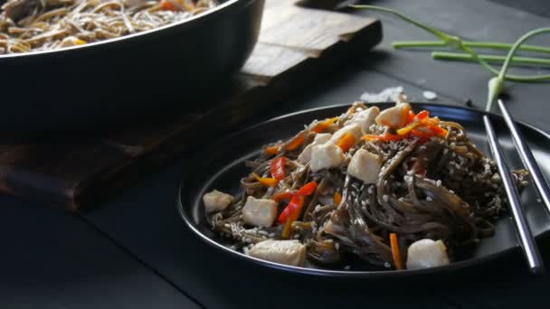 Asiatische Buchweizen-Nudeln Soba mit Gemüse Zwiebelschoten, Karotten, Paprika und Sesam und Hühnerstücken, auf einem stilvollen schwarzen Teller und chinesischen Essstäbchen im Stil der zurückhaltenden — Stockvideo