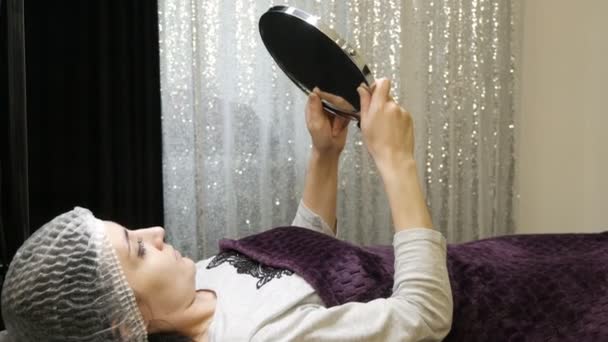 Модель в косметологической комнате лежит в медицинской шапке после процедур и смотрит в зеркало — стоковое видео