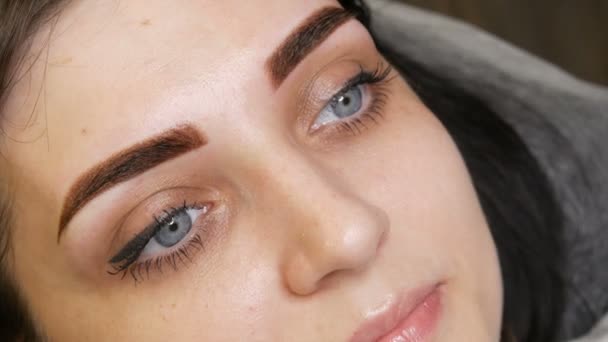 Das Gesichtsporträt einer schönen jungen Frau mit blauen Augen und langen Wimpern nach der Prozedur des Permanent Make-up durch Microblading mit Augenbrauentätowierung liegt auf einer Couch in einem Schönheitssalon — Stockvideo