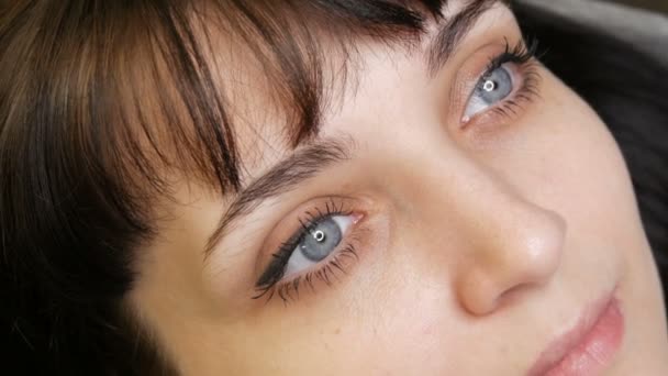 El retrato de la cara de una hermosa joven con ojos azules y pestañas largas antes del procedimiento de maquillaje permanente por microblading con tatuaje de cejas se encuentra en un sofá en un salón de belleza — Vídeos de Stock