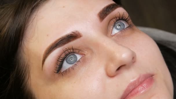 Ansiktsporträtt av en vacker ung kvinna med blå ögon och långa ögonfransar efter förfarandet för permanent make up genom microblading med ögonbrynstatuering ligger på en soffa i en skönhetssalong — Stockvideo