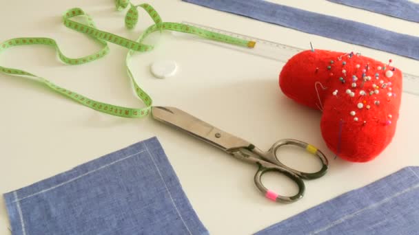 Kit de costura, tijeras de coser, trozos de tela azul, un cojín de alfiler rojo con agujas en forma de corazón, un centímetro sobre un fondo de mesa blanco. Taller de costura, flujo de trabajo de fábrica de juguetes — Vídeos de Stock