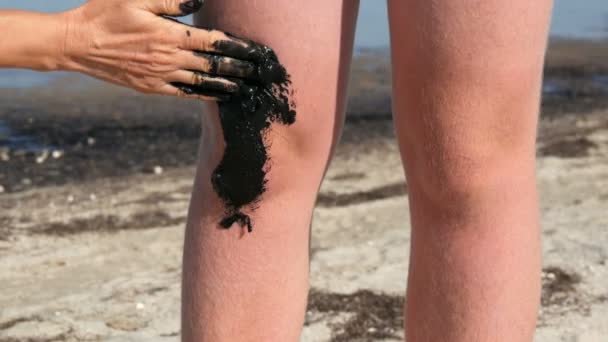 Máma ruka skvrny hojící bahno na dospívající syn nohu a koleno u jezera slané vody. Slaná černá hlína je dobrá pro lidi se zdravotními a kožními problémy. Odpočiňte si v lázních. Exfoliation scrub beauty treatment — Stock video
