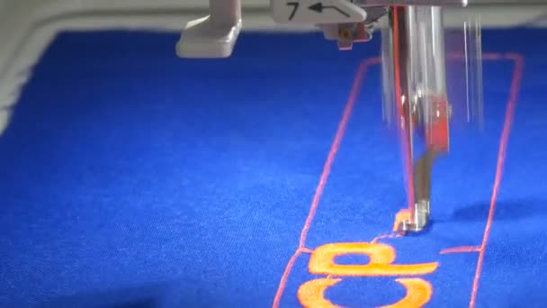 A special needle from a sewing machine embroider orange letters on blue fabric in a tailor shop or sewing workshop close up view — Stock Video