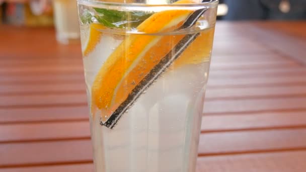 Λεμονάδα σε ποτήρι με φέτες πορτοκαλιού, λεμονιού, μέντας, με καλαμάκι για χυμό σε ξύλινο τραπέζι, καλοκαιρινό δροσιστικό ποτό — Αρχείο Βίντεο