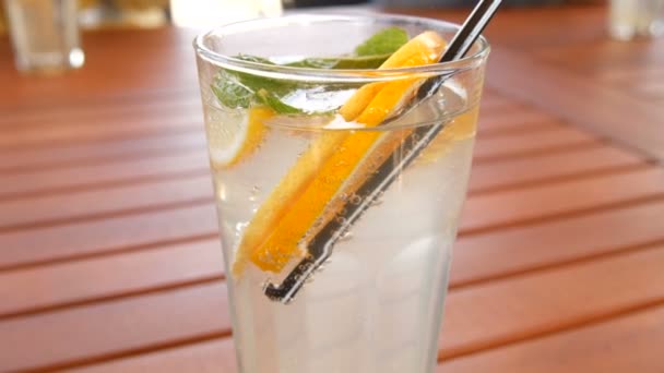 Bombillas de limonada en un vaso con rodajas de naranja, limón, menta, con una paja para el jugo en una mesa de madera, bebida refrescante de verano — Vídeo de stock