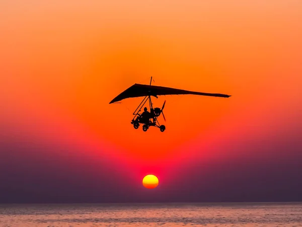 The person flying on a trike with the motor against The hang-glider is flying against the sunset.