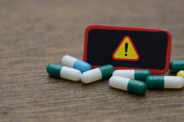 İlaç Tehlikesi: Ahşap bir yüzey üzerinde sarı uyarı işareti olan dağınık mavi ve yeşil kapsüllerin yer aldığı ilaç risklerinin kavramsal bir görüntüsü.