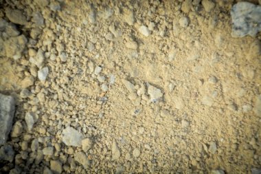 Kuru, sarımsı kahverengi bir toprak yüzeyinin makro fotoğrafı, doğal bir zemin için kum, kir ve küçük kayaların detaylı dokusunu vurguluyor..