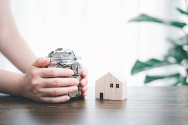 Önünde bozuk para dolu kavanoz / şişe şişesi bulunan iki el, ahşap ev modeli ve ağaç ev dekorasyonu arka planında. Finansal para tasarrufu / ev veya gayrimenkul yatırımı kavramı