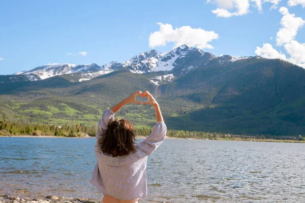 Happy woman spread her arms enjoying nature and relaxation (rear view) Brunette girl relaxes in the mountains on the lake. Young woman holding hands in the form of a heart. Travel and Vacation Concept