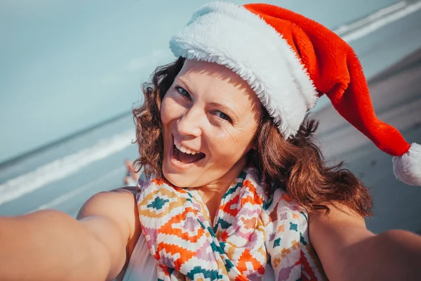 People, holidays and Christmas ( New Year) concept - young woman in Santa hat taking selfie on beach.