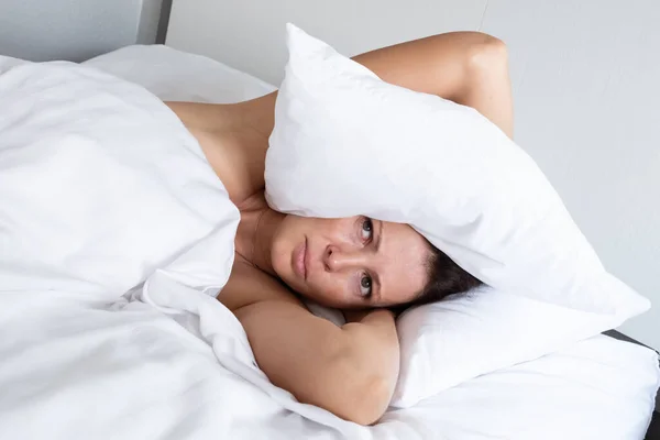 Woman suffering from noisy neighbors covers her head with a pillow. Stress and bad sleep concept