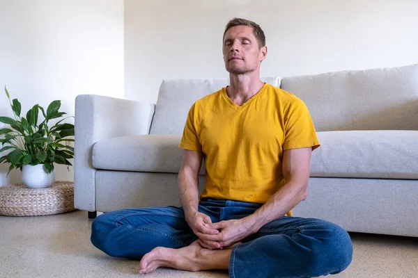 Handsome man with eyes closed sitting on floor in Lotus Pose and meditating at home