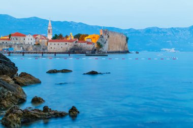 Alacakaranlıkta eski Budva şehrine, Karadağ 'daki Adriyatik sahil şeridine, öndeki büyük taşlara, mavi saate bakın..
