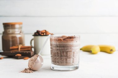 Çikolatalı protein içeceği, muzlu beyaz masada milkshake smoothie, ölçü kaşığında protein tozu, protein barı, badem fıstığı ve tarçın çubukları..