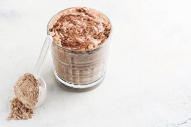 Çikolatalı protein içeceği, beyaz masada süt tozu ve ölçü kaşığında protein tozu..