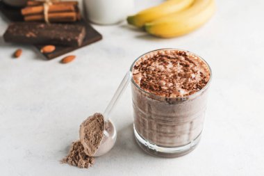 Çikolatalı protein içeceği, muzlu beyaz masada milkshake smoothie, ölçü kaşığında protein tozu, protein barı, badem fıstığı ve tarçın çubukları..
