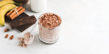 Çikolatalı protein içeceği, muzlu beyaz masada milkshake smoothie, ölçü kaşığında protein tozu, protein barı, badem fıstığı ve tarçın çubukları..