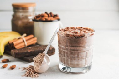 Çikolatalı protein içeceği, muzlu beyaz masada milkshake smoothie, ölçü kaşığında protein tozu, protein barı, badem fıstığı ve tarçın çubukları..