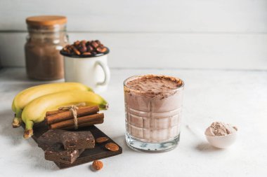 Çikolatalı protein içeceği, muzlu beyaz masada milkshake smoothie, ölçü kaşığında protein tozu, protein barı, badem fıstığı ve tarçın çubukları..