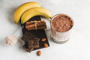 Çikolatalı protein içeceği, muzlu beyaz masada milkshake smoothie, ölçü kaşığında protein tozu, protein barı, badem fıstığı ve tarçın çubukları..