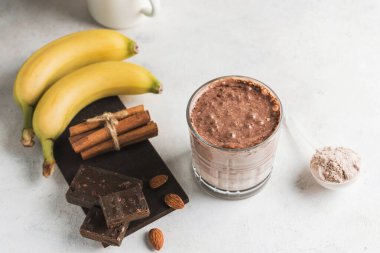 Çikolatalı protein içeceği, muzlu beyaz masada milkshake smoothie, ölçü kaşığında protein tozu, protein barı, badem fıstığı ve tarçın çubukları..