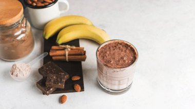 Çikolatalı protein içeceği, muzlu beyaz masada milkshake smoothie, ölçü kaşığında protein tozu, protein barı, badem fıstığı ve tarçın çubukları..
