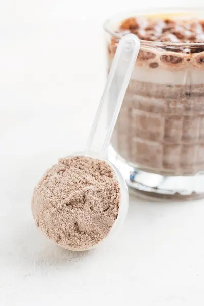 Whey protein powder in measuring spoon with glass of chocolate protein drink, milkshake smoothie on white table.