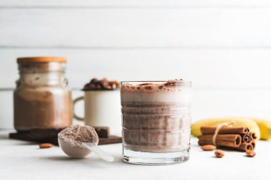 Çikolatalı protein içeceği, muzlu beyaz masada milkshake smoothie, ölçü kaşığında protein tozu, protein barı, badem fıstığı ve tarçın çubukları..