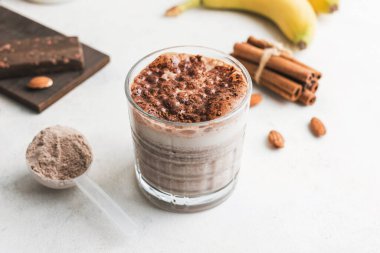Çikolatalı protein içeceği, muzlu beyaz masada milkshake smoothie, ölçü kaşığında protein tozu, protein barı, badem fıstığı ve tarçın çubukları..