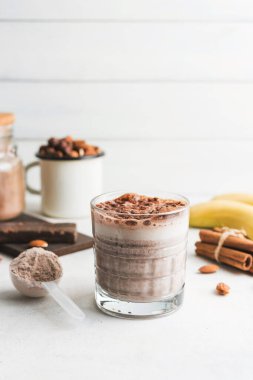 Çikolatalı protein içeceği, muzlu beyaz masada milkshake smoothie, ölçü kaşığında protein tozu, protein barı, badem fıstığı ve tarçın çubukları..