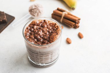 Çikolatalı protein içeceği, muzlu beyaz masada milkshake smoothie, ölçü kaşığında protein tozu, protein barı, badem fıstığı ve tarçın çubukları..