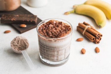 Çikolatalı protein içeceği, muzlu beyaz masada milkshake smoothie, ölçü kaşığında protein tozu, protein barı, badem fıstığı ve tarçın çubukları..