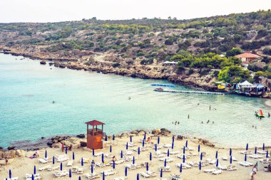 Protaras, Kıbrıs - 16 Eylül 2018: Gün ışığı günbatımı manzaralı Konnos Plajı'na kum üzerinde rahatlatıcı ve suda yüzen insanlar. Su ulaştırma araçları yola yakın kira. Arka planda dağlar