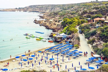 Protaras, Kıbrıs - 16 Eylül 2018: Gün ışığı günbatımı manzaralı Konnos Plajı'na kum üzerinde rahatlatıcı ve suda yüzen insanlar. Su ulaştırma araçları yola yakın kira. Arka planda dağlar