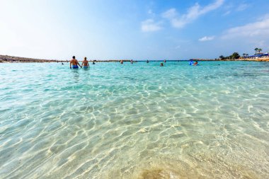 Nissi Beach yaz görünümüne renkli parlak mavi su ve gökyüzü bulutlu. Yüzme ve kum üzerinde rahatlatıcı turistler. Negatif kopya alanı, metin için yer. Ayia Napa, Kıbrıs
