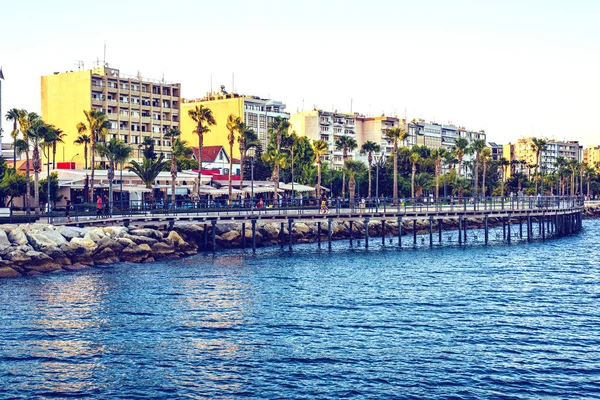 Sunset view to city and water. People walking on promenade alley and relaxing in restaurants. Sun reflecting on buildings facade. Negative copy space, place for text. Limassol, Cyprus