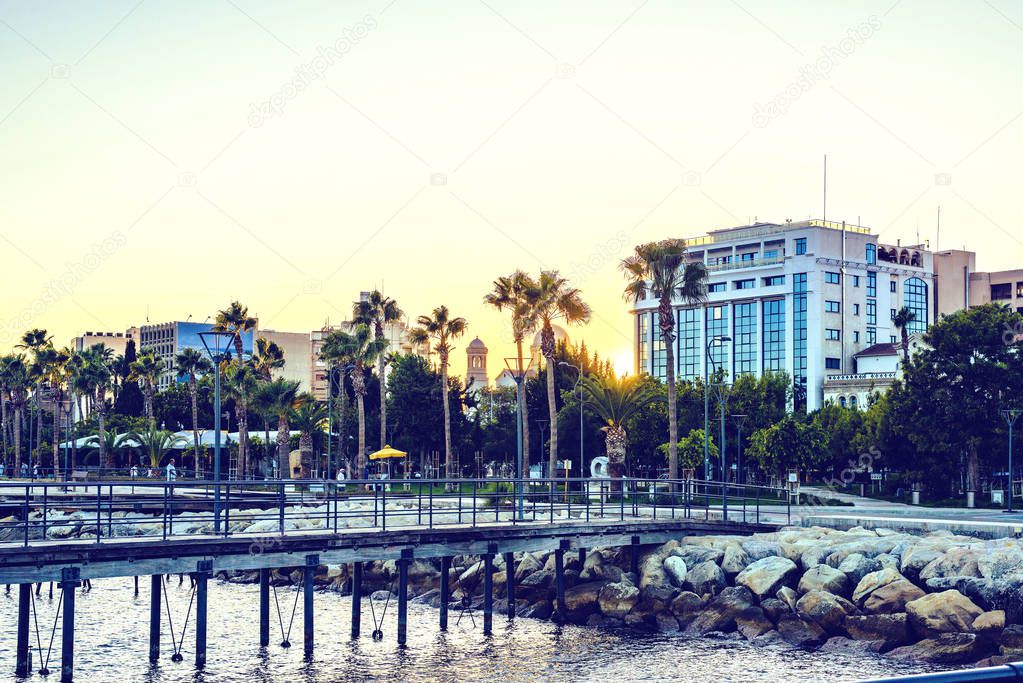 Sunset view to city and water. People walking on promenade alley and relaxing in restaurants. Sun reflecting on buildings facade. Negative copy space, place for text. Limassol, Cyprus