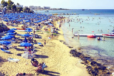 Protaras, Kıbrıs - 23 Eylül 2017: Gün ışığı plaj su rahatlatıcı ve sahil üzerinde güneşlenme insanlarla görünümüne. Yemyeşil ağaçlar ve arka plan üzerinde şehir. Negatif kopya alanı, metin için yer