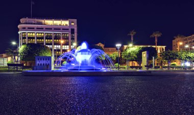 Gece manzara mavi ışık parlayan ile modern kamu çeşme. Otel ve park arka plan üzerinde. Negatif kopya alanı, metin için yer. Molos Promenade, Limassol, Kıbrıs