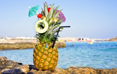Ananas meyve ve mini bir şemsiye ile harika egzotik bir tarzda dekore edilmiştir. Taşlar ve deniz suyu arka plan üzerinde. Negatif kopya alanı, metin için yer. Nissi Beach, Ayia Napa, Kıbrıs.