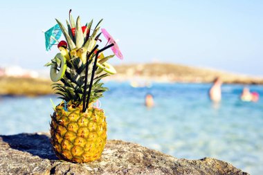 Ananas meyve ve mini bir şemsiye ile harika egzotik bir tarzda dekore edilmiştir. Taşlar ve deniz suyu arka plan üzerinde. Negatif kopya alanı, metin için yer. Nissi Beach, Ayia Napa, Kıbrıs.
