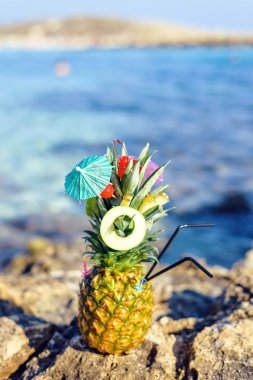 Ananas meyve ve mini bir şemsiye ile harika egzotik bir tarzda dekore edilmiştir. Taşlar ve zemin üzerine mavi deniz. Negatif kopya alanı, metin için yer. Nissi Beach, Ayia Napa, Kıbrıs.