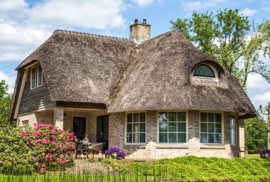 Güneşli bir günde rustik thatched çatı ile geleneksel ev. Ağaçlar, çiçekler ve parlak mavi gökyüzü bulutlu arka plan üzerinde. Giethoorn, Hollanda
